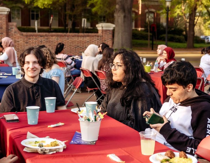 Three people sit at an outdoor table with food and drinks during a communal gathering.