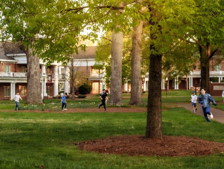 Several people walk and run across a green campus lawn surrounded by trees and buildings.