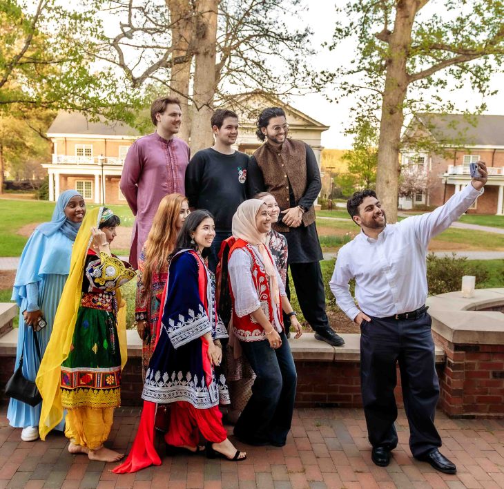 A group of people gather outdoors as one person takes a selfie with everyone smiling and posing behind them.