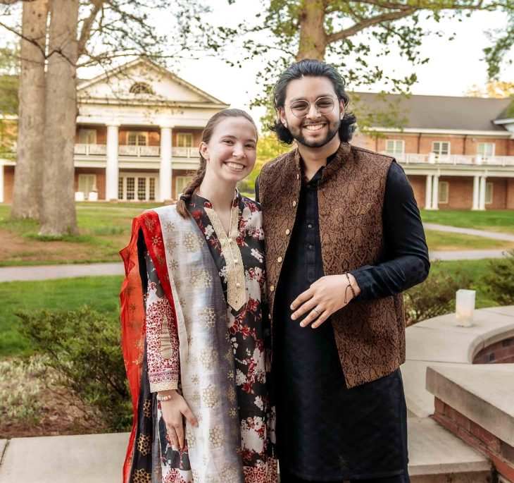 A smiling pair in traditional clothing stand together outdoors on a campus lawn with a columned building behind them.
