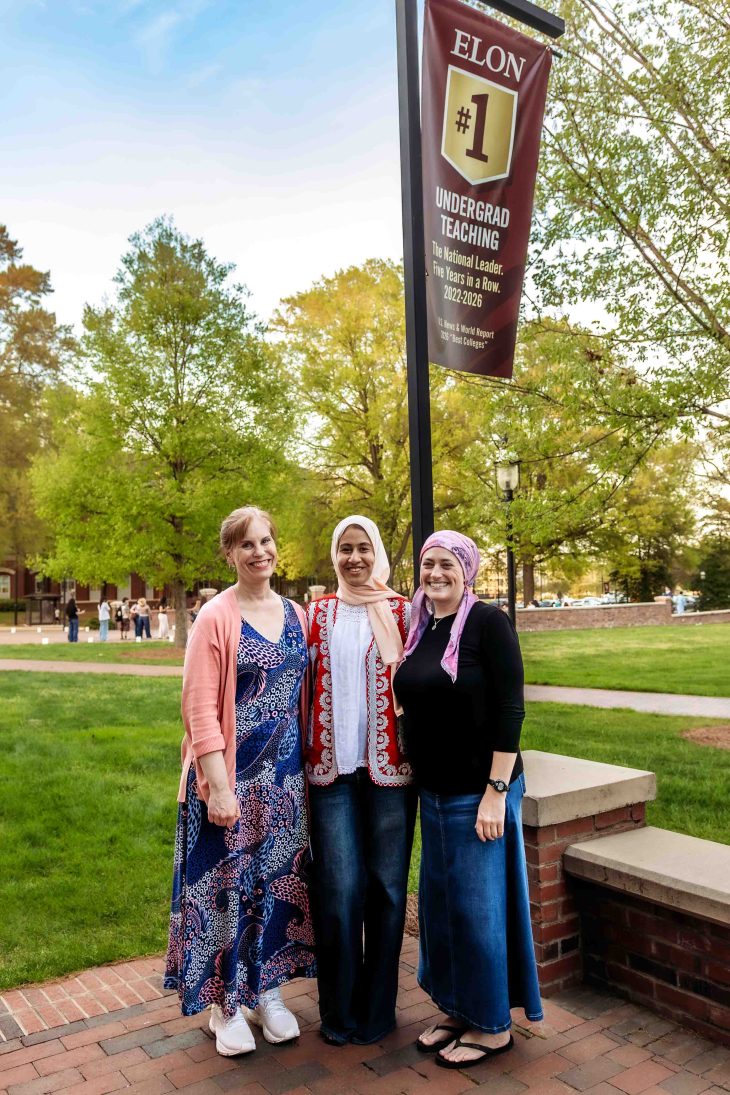 Three women stand together smiling beneath a campus banner that reads “Elon #1 Undergrad Teaching.”