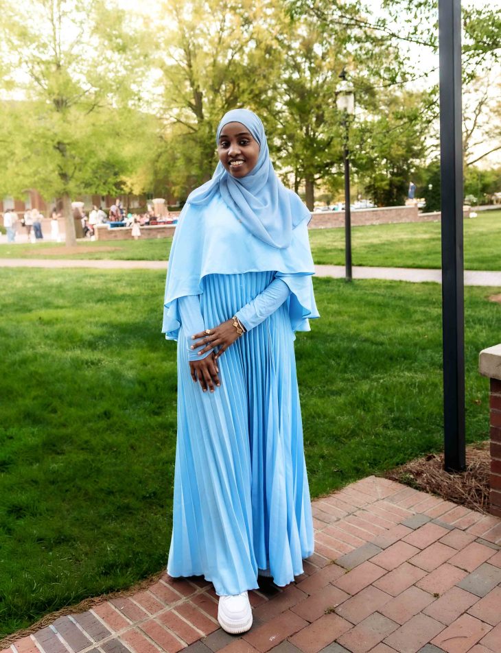 A woman in a flowing light blue dress and matching hijab stands on a brick path in a grassy campus setting.