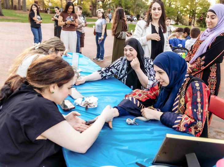 A group of women gather around a table outdoors, engaging in an activity while others stand and watch nearby.