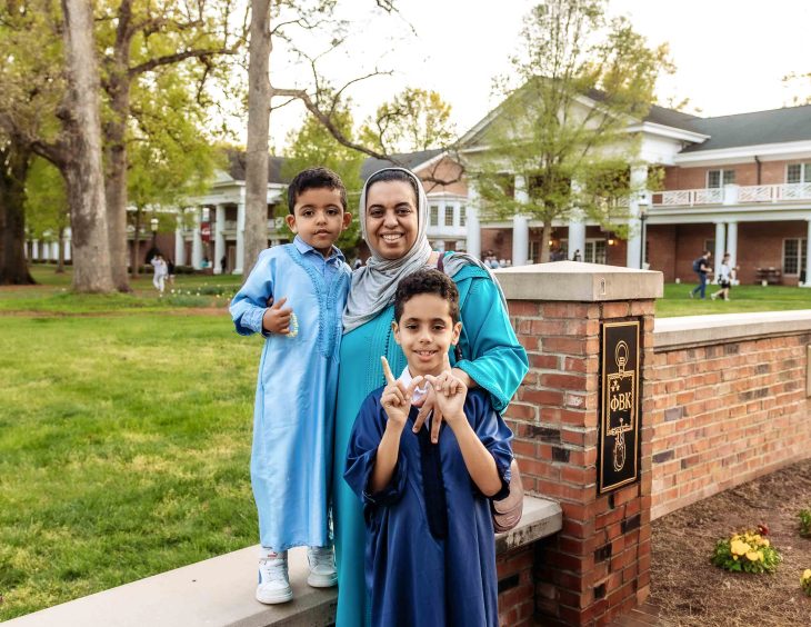 A woman wearing a hijab poses with two young boys on a campus walkway, all smiling at the camera.