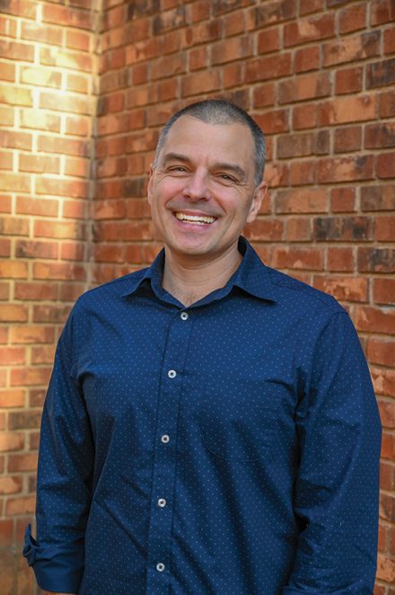 A man smiles while standing outdoors in front of a red brick wall, wearing a blue button-down shirt.