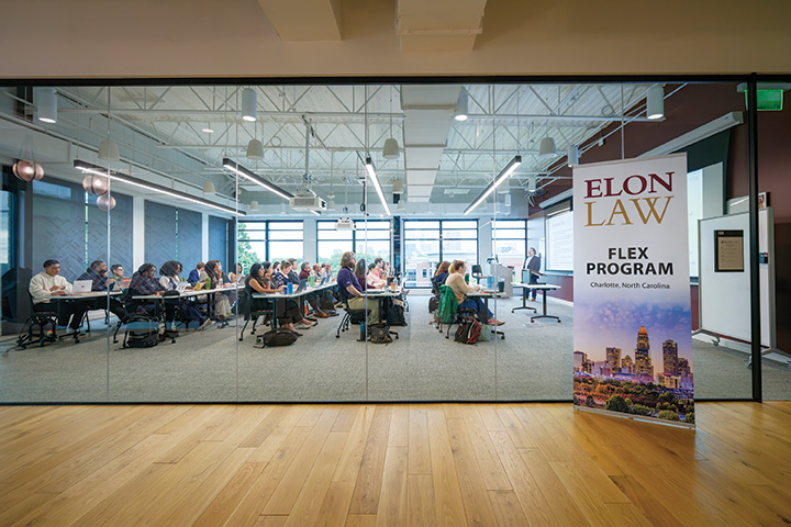 Students sit in a modern classroom behind a glass wall as an instructor teaches, with a sign reading “Elon Law Flex Program, Charlotte, North Carolina” in the foreground.