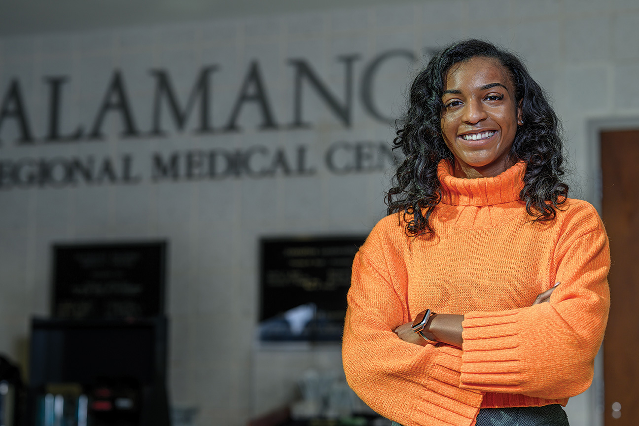 A woman smiles with arms crossed while standing indoors in front of a wall with “Alamance Regional Medical Center” signage.