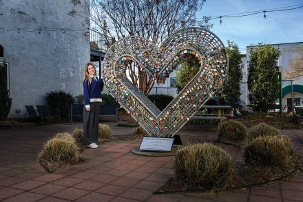 A woman stands beside a large heart-shaped sculpture covered in colorful locks in an outdoor courtyard.