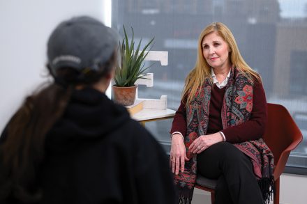 A woman sits facing another person during a counseling session in a bright office, with a plant and decorative letter “E” on a nearby table.