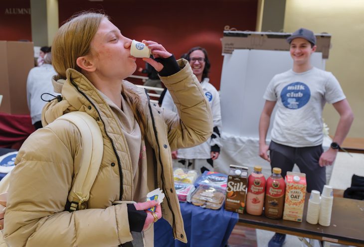 A student samples a small cup of milk at a campus table staffed by a student “milk club,” with cartons of almond, oat and flavored milk displayed and other students smiling in the background.