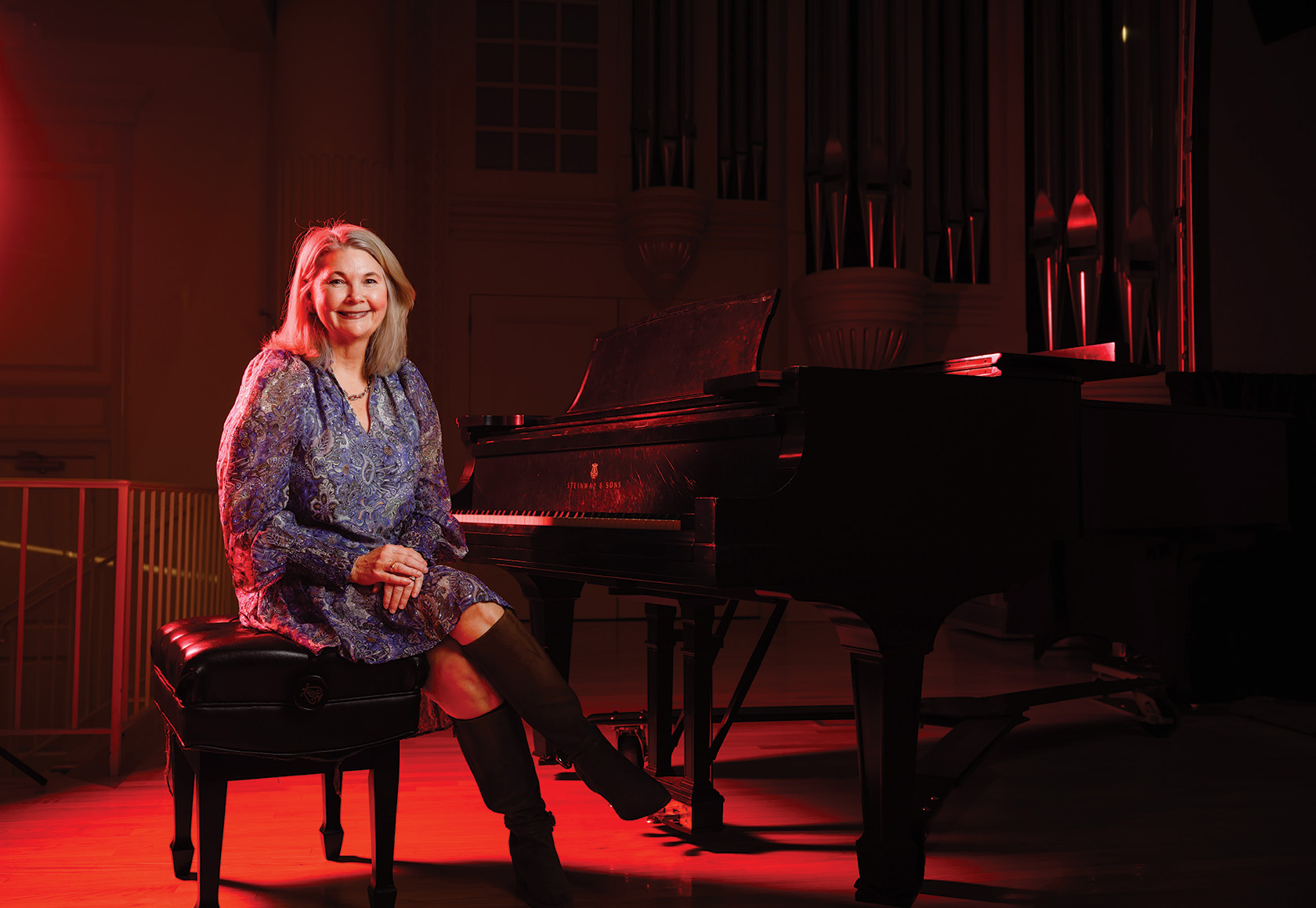A woman sits beside a grand piano on a concert stage, lit with warm red lighting, with organ pipes visible in the background.