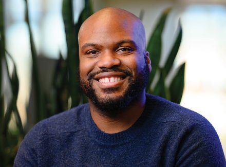 A man with a beard smiles in a close-up portrait, seated indoors with green plants softly blurred in the background.