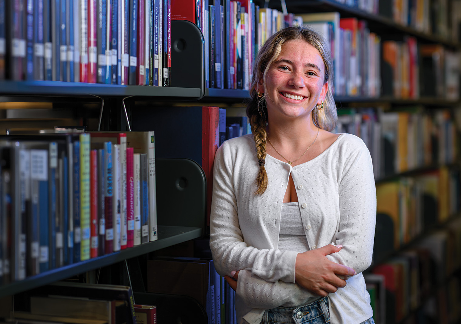 A student smiles while standing between bookshelves in a library, arms folded, with rows of colorful books in the background.