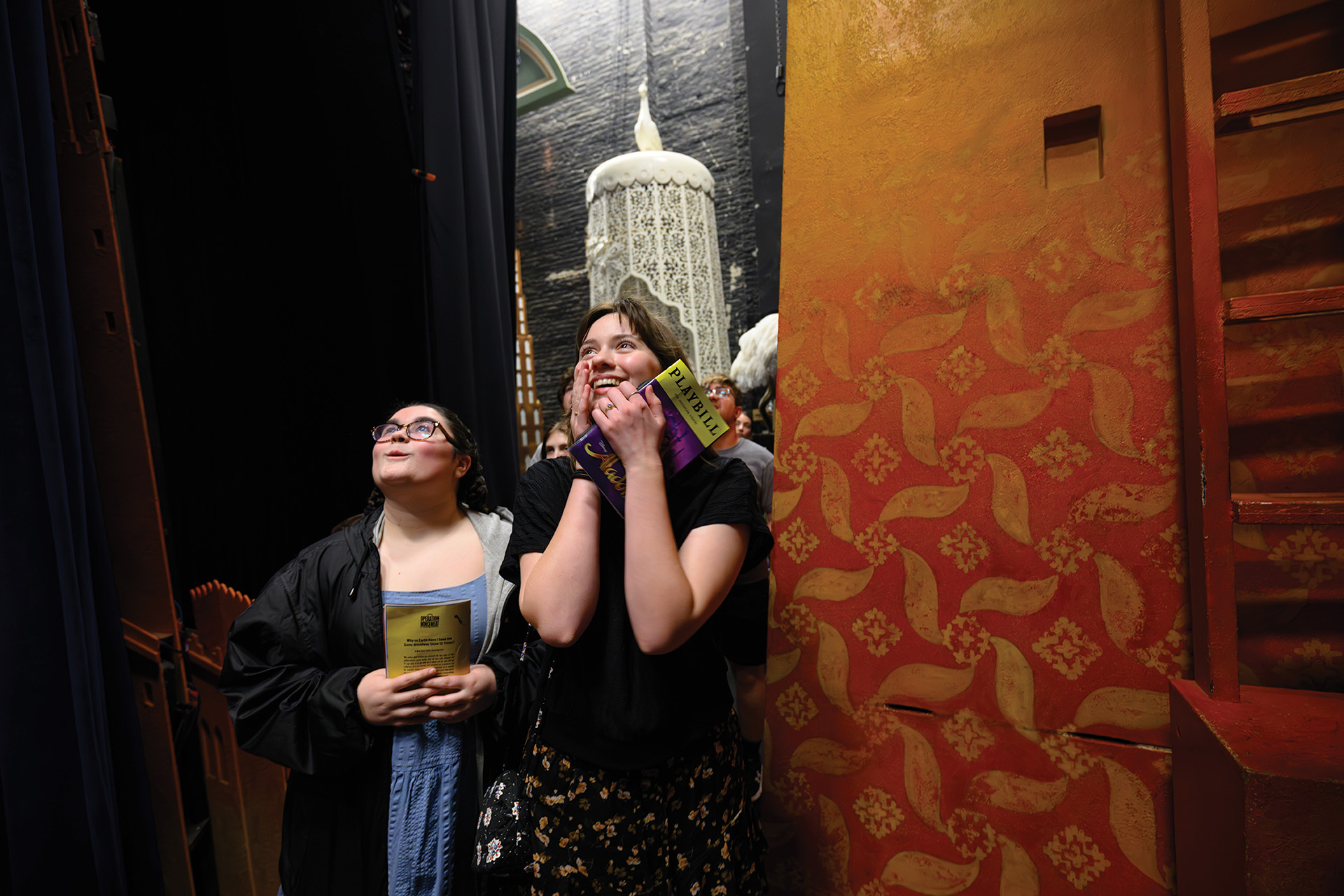 Students stand backstage in a theater, looking upward in awe while holding programs beside colorful set pieces.