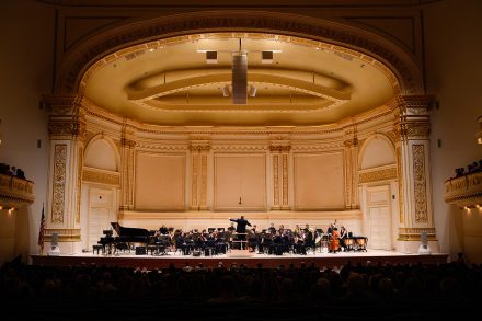 A wind ensemble performs onstage in an ornate concert hall, with a conductor leading musicians before an audience.