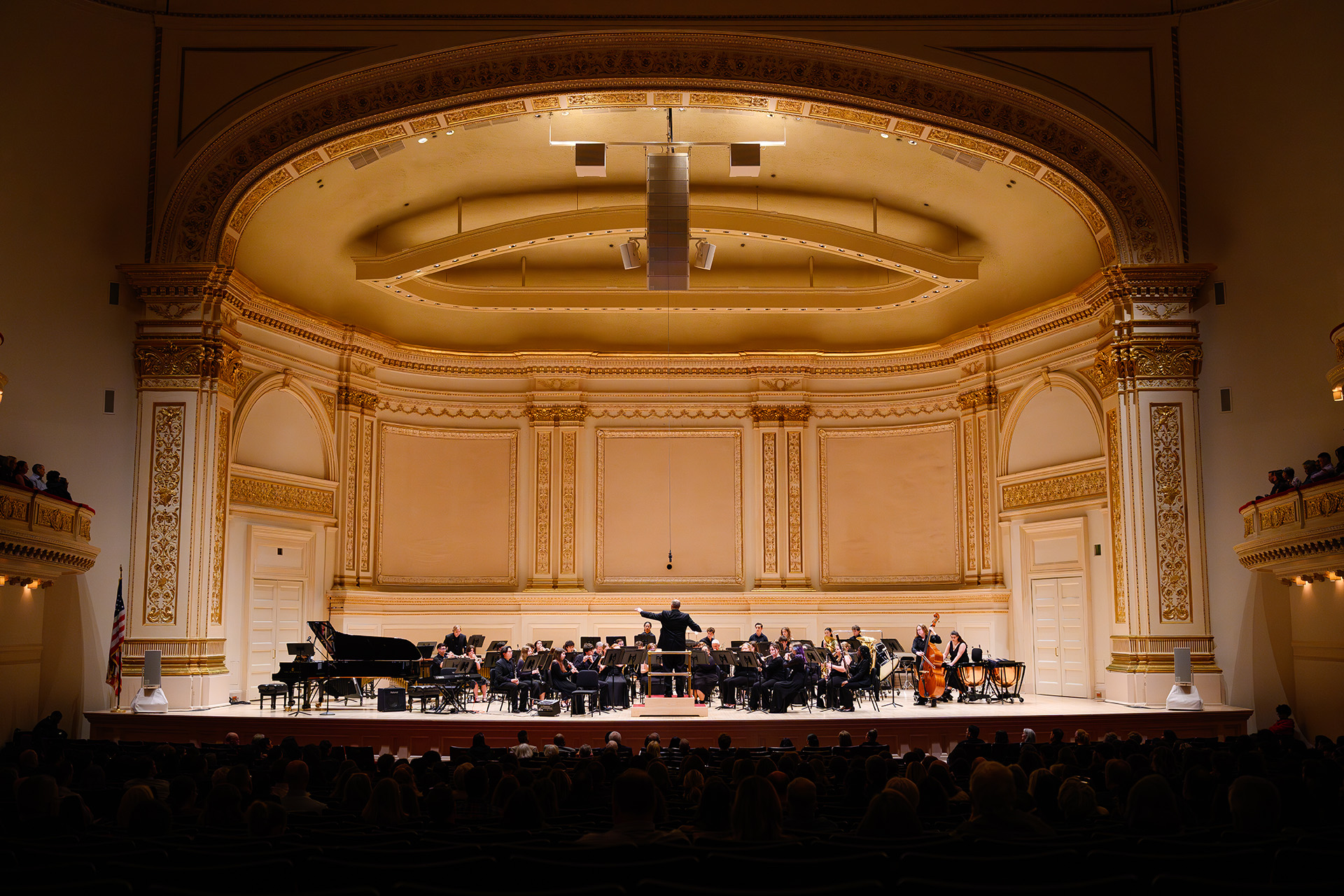 A wind ensemble performs onstage in an ornate concert hall, with a conductor leading musicians before an audience.