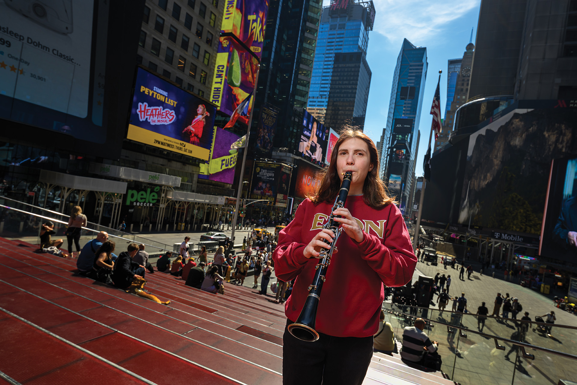 A student wearing an Elon sweatshirt plays a clarinet in Times Square, surrounded by bright billboards and city crowds.