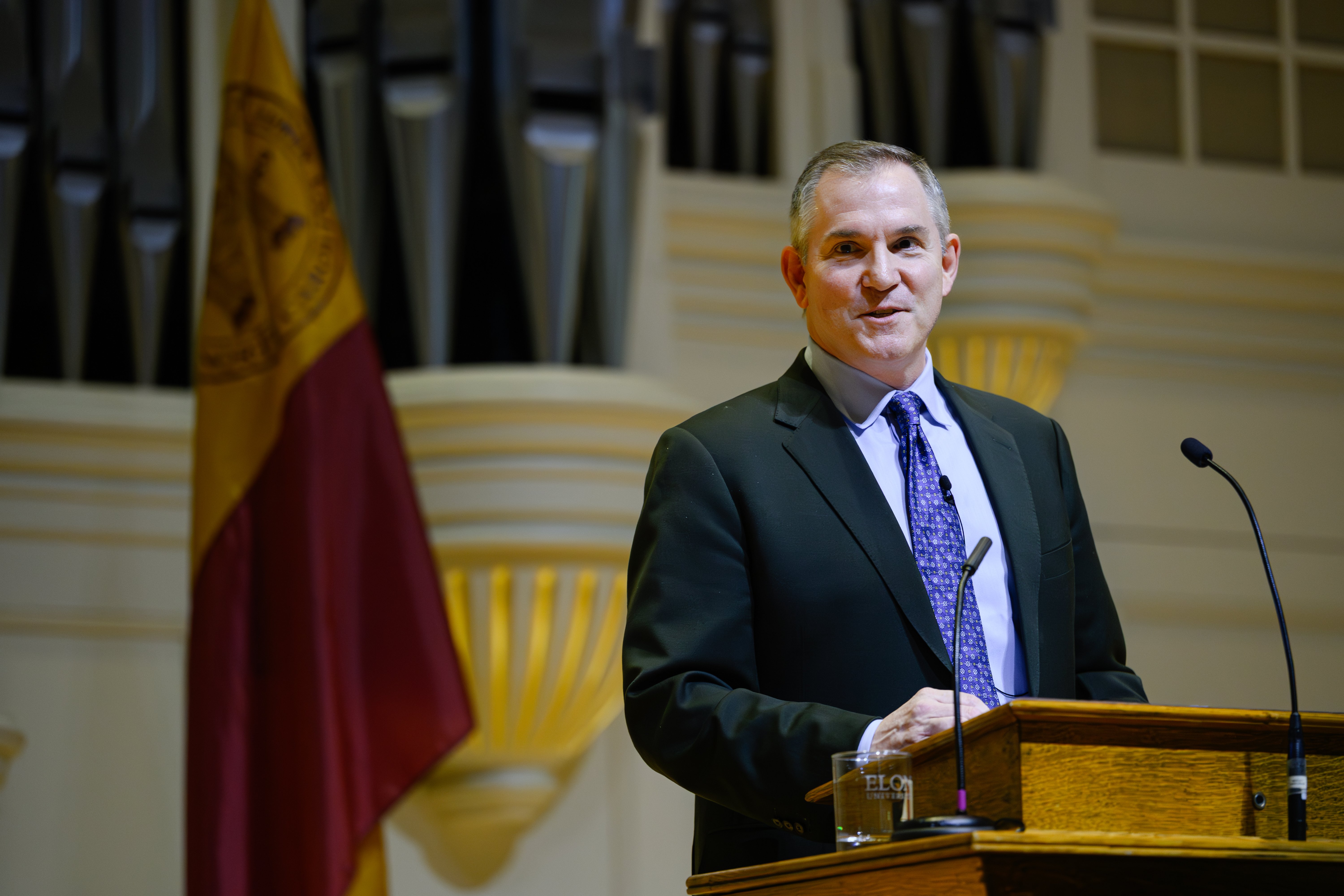 A man in a suit stands at a podium delivering a talk in an auditorium with flags in the background.