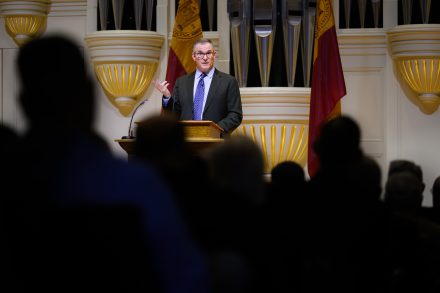 A person stands at a podium speaking to an audience. Behind them, pipes and flags displaying the Harvard University emblem are visible.