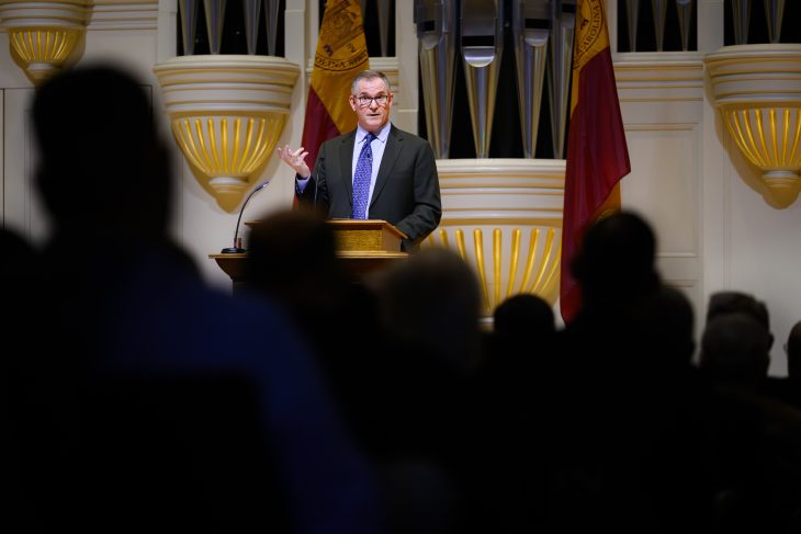 A person stands at a podium speaking to an audience. Behind them, pipes and flags displaying the Harvard University emblem are visible.