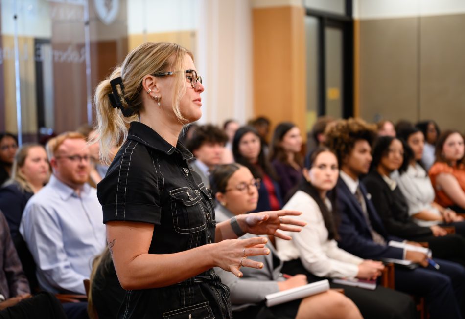 A woman in professional attire stands among classmates ina. crowded courtroom.