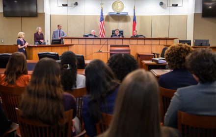 A view from the back of a full courtroom, with three judges in black robes on the bench, and two judges in professional attire with a law professor standing to the side.