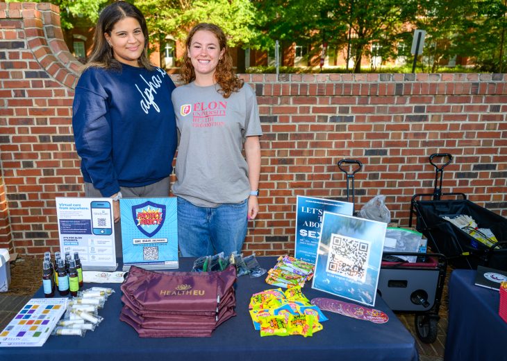 Two student staff members stand behind a HealthEU resource table, sharing wellness information, giveaways and campus resources with the community.