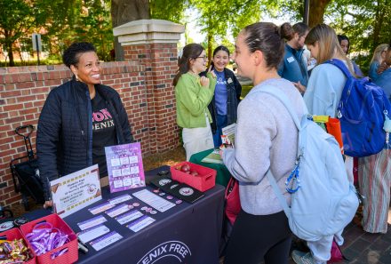 A staff member shares information about recovery resources with a student at a campus resource table, while others connect and explore materials nearby.