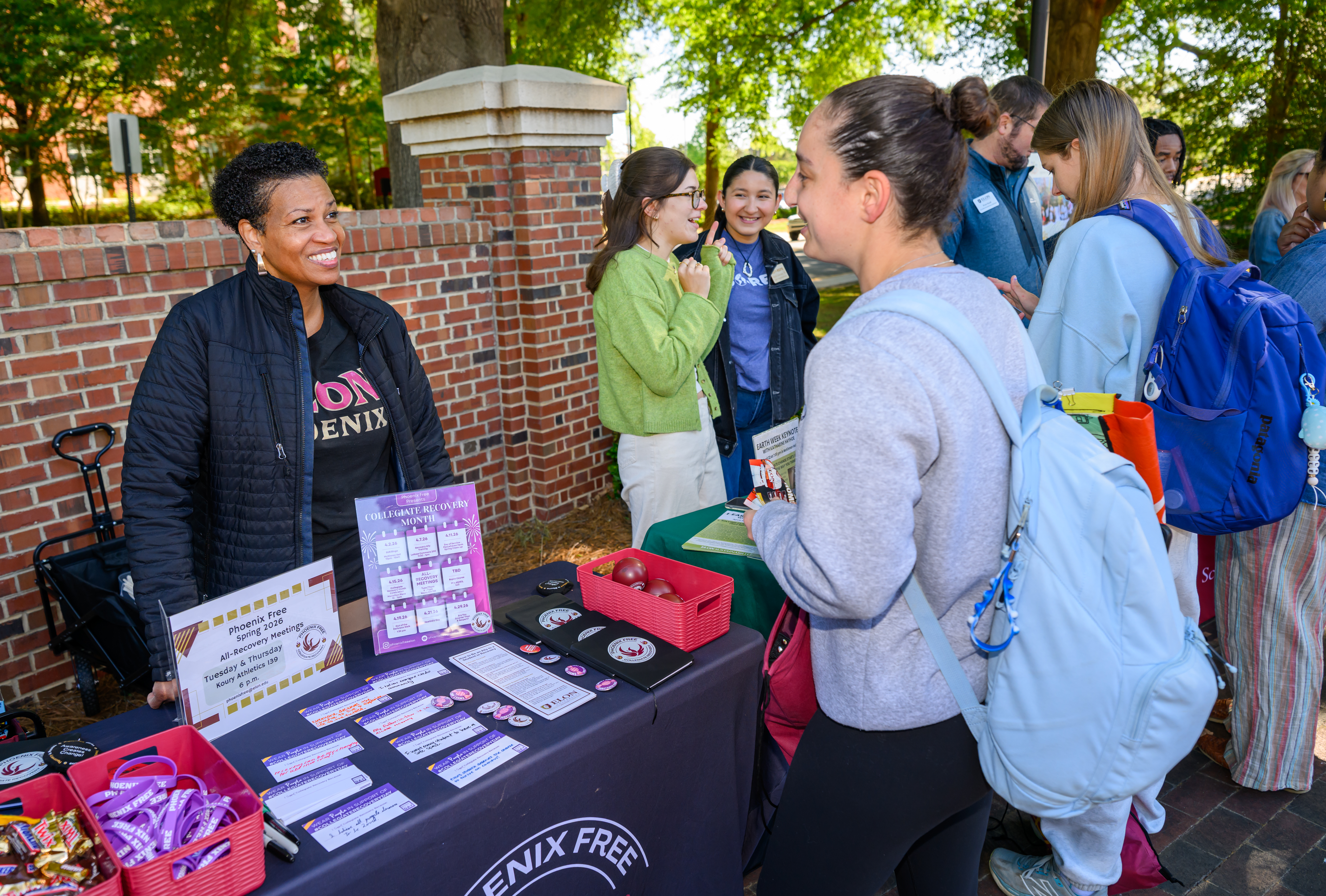 A staff member shares information about recovery resources with a student at a campus resource table, while others connect and explore materials nearby.