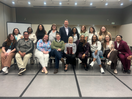 A group of conference attendees poses together in a meeting room, with one individual seated at the center holding an award plaque while colleagues stand and sit around them, smiling at the camera.