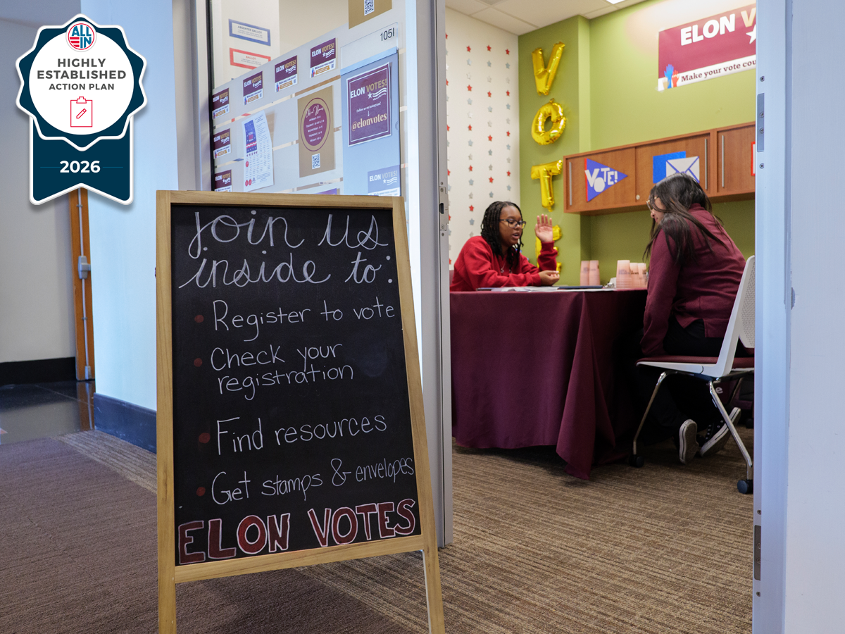Chalkboard sign reading “Join us inside to: register to vote, check your registration, find resources, get stamps & envelopes” stands outside a campus office where two students talk at a voter engagement table; “Elon Votes” and an ALL IN 2026 Highly Established Action Plan badge are visible.