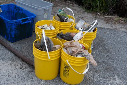 Yellow buckets filled with collected trash—plastic bottles, cans, and debris—sit on the ground after a cleanup effort.
