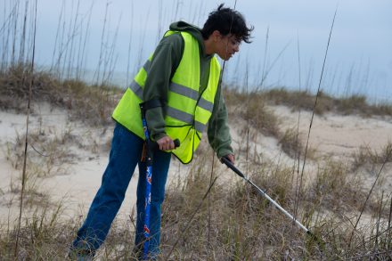 Student wearing a safety vest uses a grabber tool to pick up litter among beach grasses near the shoreline.