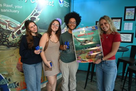 Four students smile while holding educational materials and a poster for Gray’s Reef National Marine Sanctuary inside a marine science exhibit space.