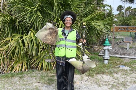 Nivea Millner '26 wearing a reflective vest smiles while holding up pieces of metal debris collected during a cleanup effort, standing in front of dense greenery.