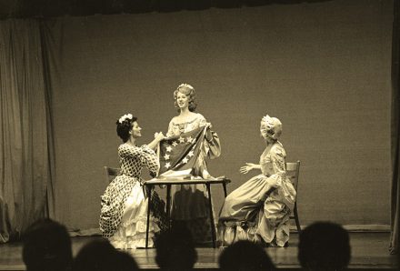 In a black-and-white archival photo, three performers in period costumes gather around a table onstage, holding a flag during a theatrical production.