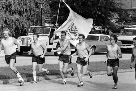 In a black-and-white archival photo, a group of runners jog along a street carrying a flag marked “American Bicentennial 1776–1976,” with cars and trees in the background.