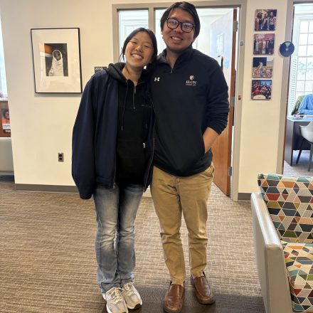 Two people stand side by side indoors, smiling with their arms around each other in an office or campus space.