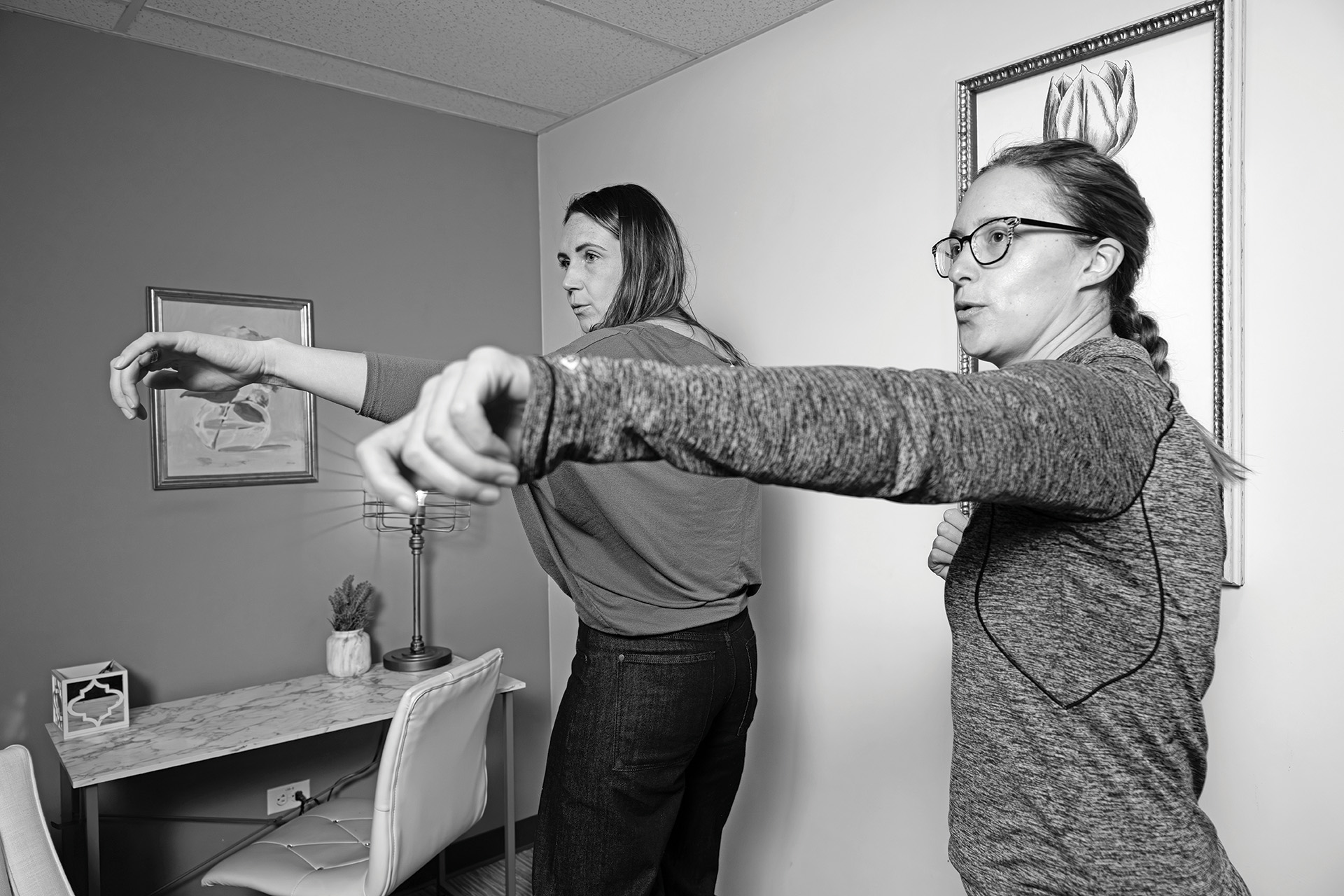 Two women extend their arms forward during a somatic boxing demonstration in an office setting, with a desk and wall art in the background.