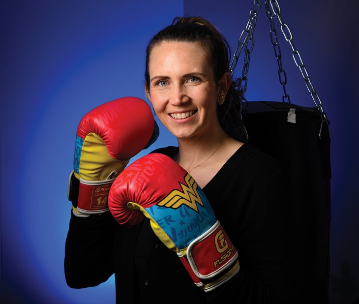 A woman smiles while wearing boxing gloves and posing beside a hanging punching bag in a studio setting.