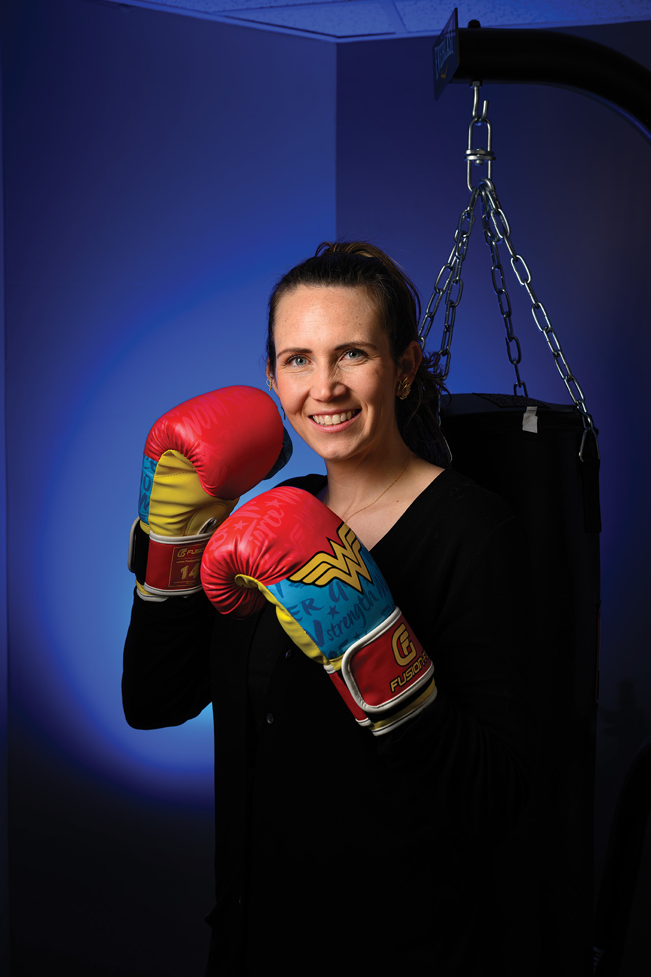 A woman smiles while wearing boxing gloves and posing beside a hanging punching bag in a studio setting.