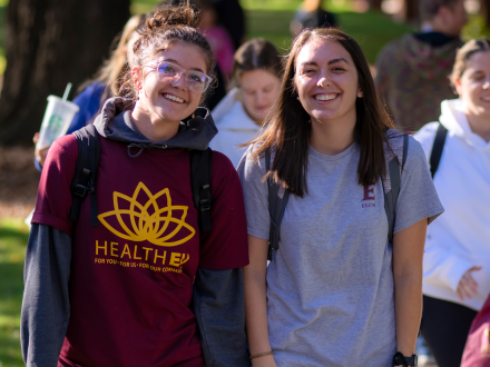 Two students walk side by side on a sunny campus path, smiling and wearing backpacks—one in a maroon HealthEU T-shirt and the other in a gray ������Ƶ T-shirt—while other students move through the background.