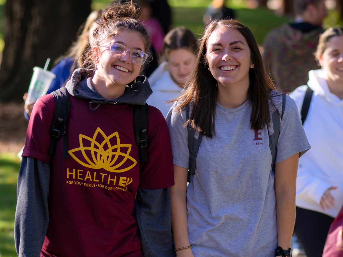 Two students walk side by side on a sunny campus path, smiling and wearing backpacks—one in a maroon HealthEU T-shirt and the other in a gray ���ϲ����� T-shirt—while other students move through the background.