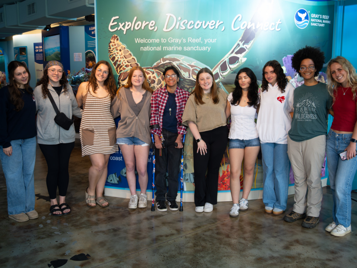 A group of students poses together inside Gray’s Reef National Marine Sanctuary’s visitor center, standing in front of a colorful exhibit that highlights marine life and coastal conservation.