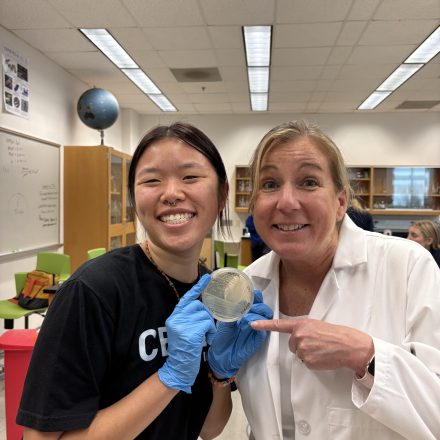 A student and instructor smile while holding a petri dish in a science classroom, with the instructor pointing to the sample.