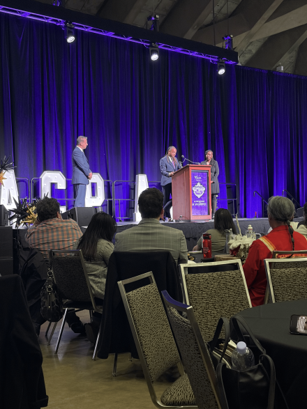 Attendees sit at round tables facing a stage where speakers stand at a podium under purple lighting, presenting during a conference session.