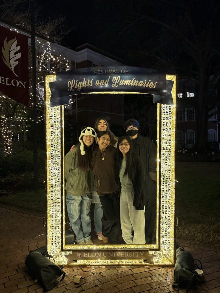 Four students pose together inside a glowing frame at a “Festival of Lights and Luminaries” event at Elon University at night.