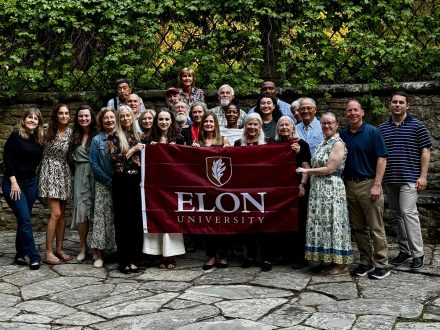 A group of people pose for a photo in front of lush greenery holding an Elon University flag