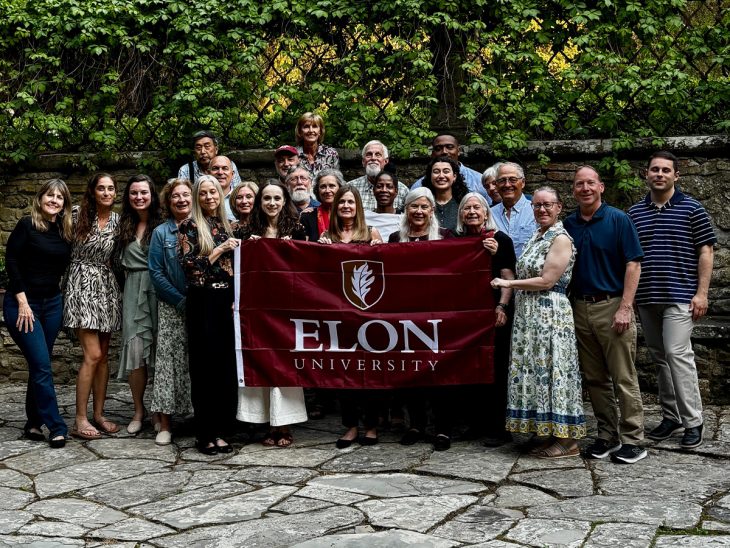 A group of people pose for a photo in front of lush greenery holding an Elon University flag