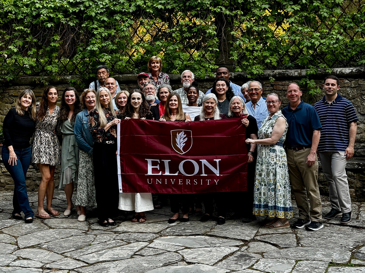 A group of people pose for a photo in front of lush greenery holding an Elon University flag